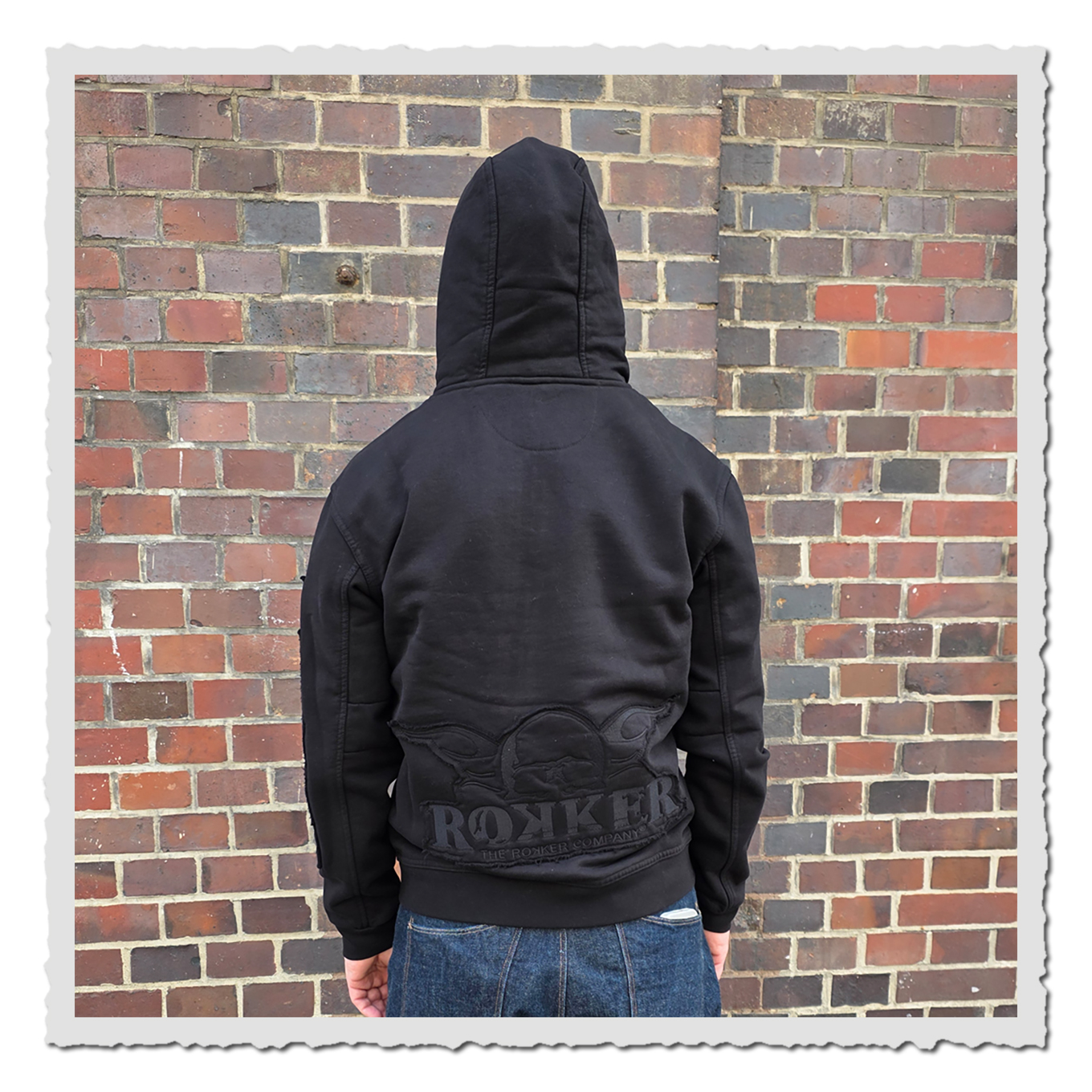 Rückansicht einer Person mit schwarzem Kapuzenhoodie vor einer rotbraunen Backsteinwand im Außenbereich.